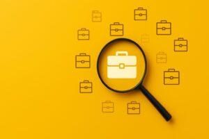 Job search concept with a magnifying glass focusing on a briefcase icon against a bright yellow background. Represents employment, career opportunities, and recruitment.