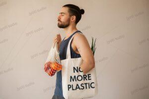 Young man holding mesh grocery bag and eco tote with No More Plastic text, sustainable shopping lifestyle concept with reusable produce bags, zero waste message and environmental awareness campaign